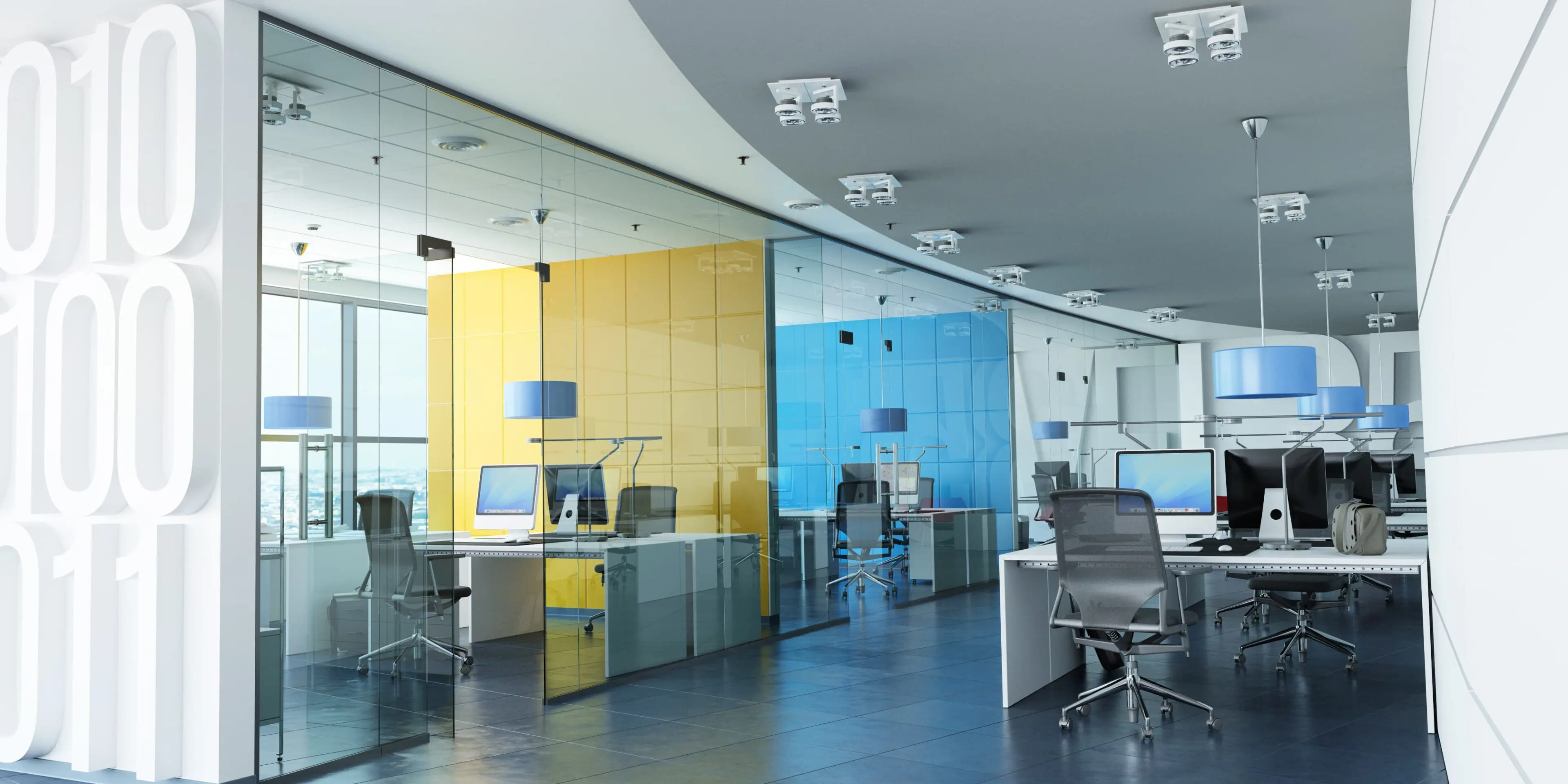 Modern office with glass walls, colorful partitions, and ergonomic chairs. Desks are equipped with computers and pendant lights hang from the ceiling.