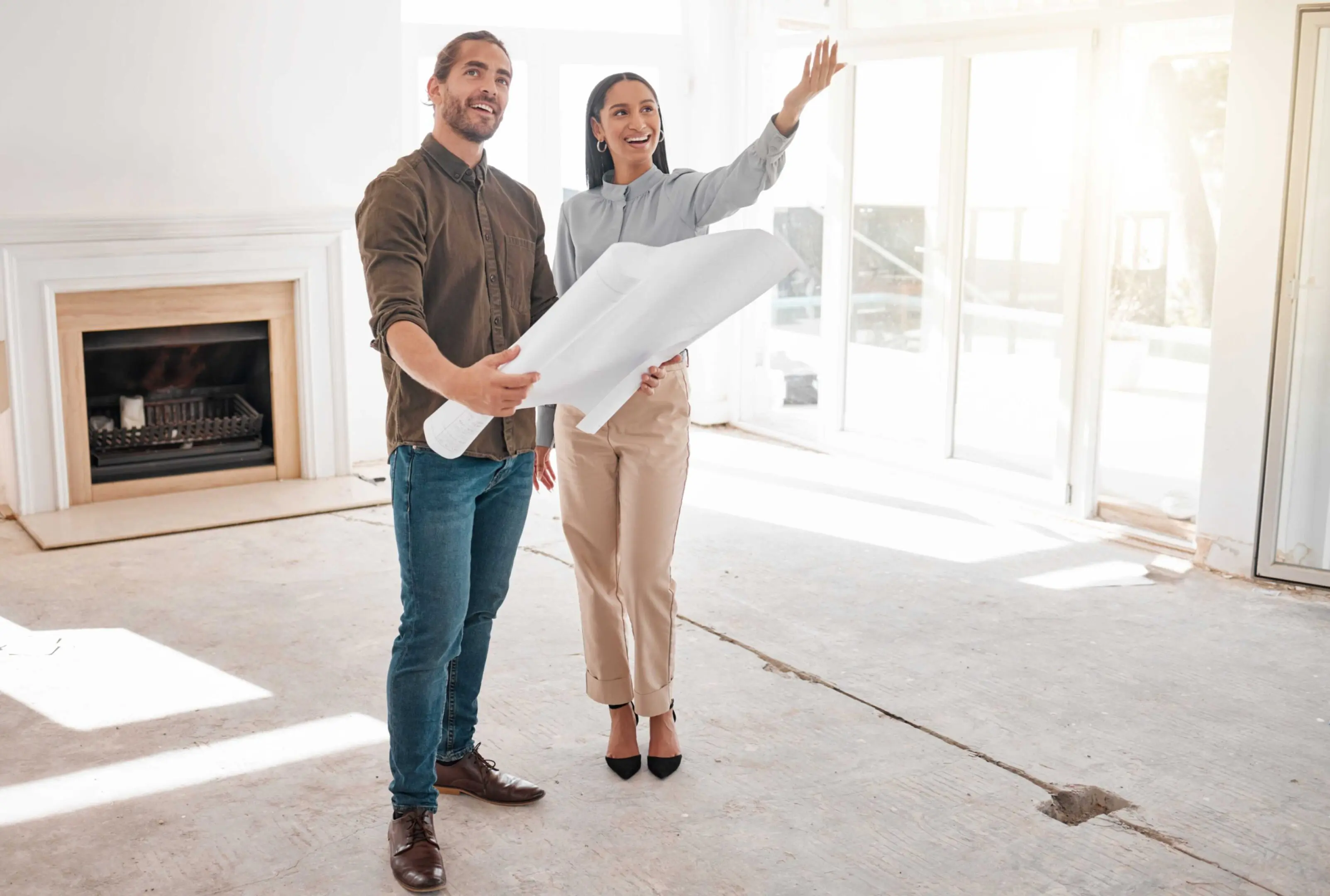 Two people stand in a room under renovation, holding blueprints and gesturing towards the windows, with an empty fireplace in the background.