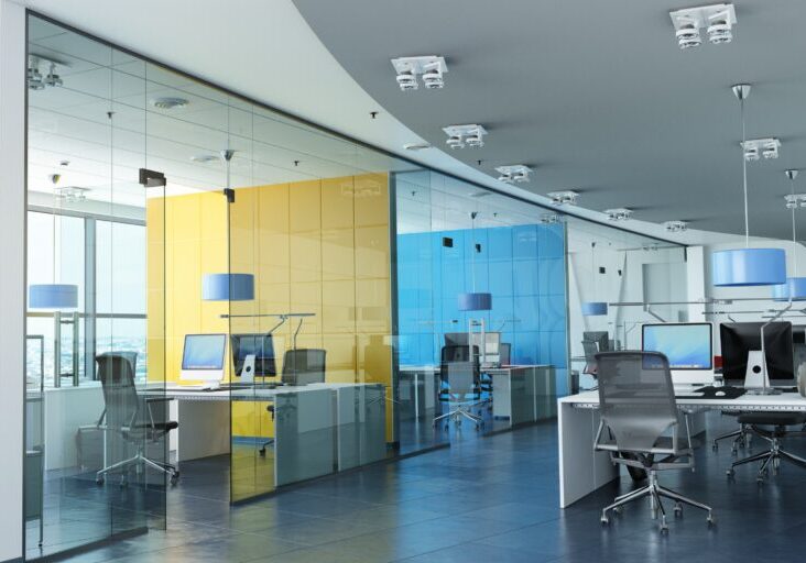 Modern office with glass walls, colorful partitions, and ergonomic chairs. Desks are equipped with computers and pendant lights hang from the ceiling.