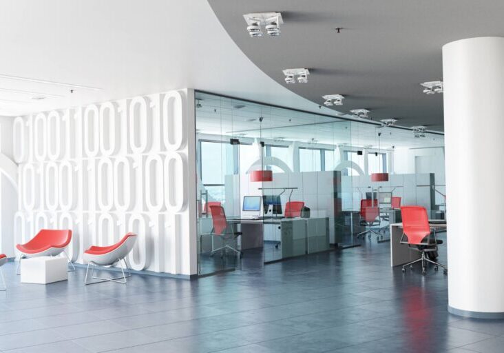 Modern office space with white walls, glass partitions, and red chairs. Open layout includes desks, computers, and ceiling lights. Large windows offer natural light.