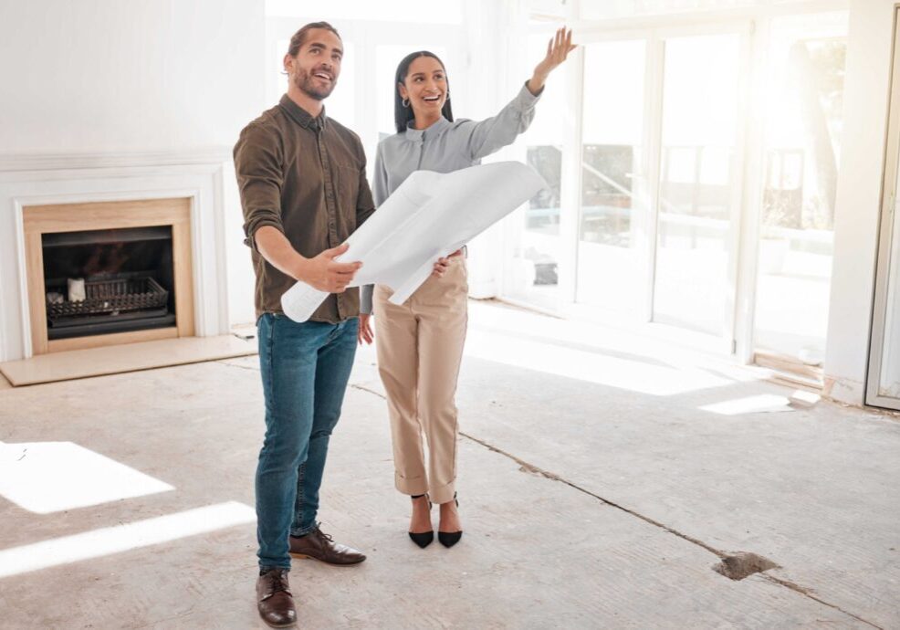 Two people stand in a room under renovation, holding blueprints and gesturing towards the windows, with an empty fireplace in the background.